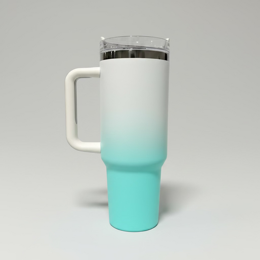 A tall handled travel tumbler with a white-to-turquoise ombre finish, a clear lid with a black rim, and a white handle, standing on a dark gray surface against a light gray background.