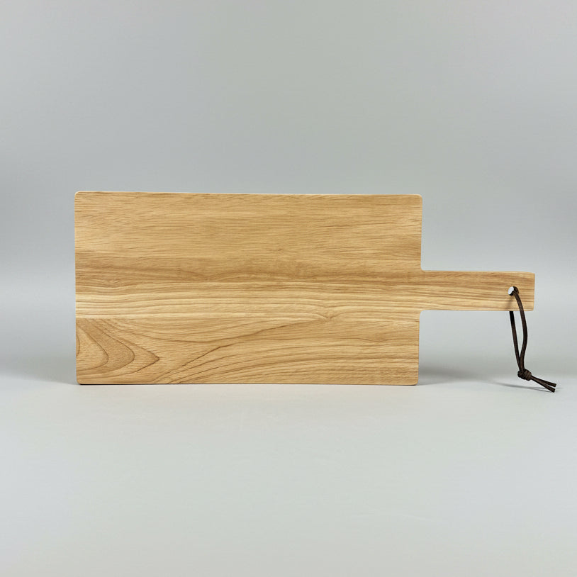 A rectangular wooden cutting board with a handle and a black hanging loop, photographed on a dark tabletop in a workshop setting.