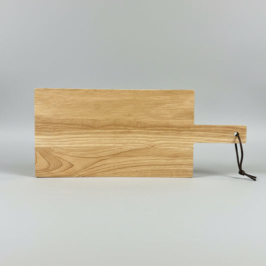 A rectangular wooden cutting board with a handle and a black hanging loop, photographed on a dark tabletop in a workshop setting.